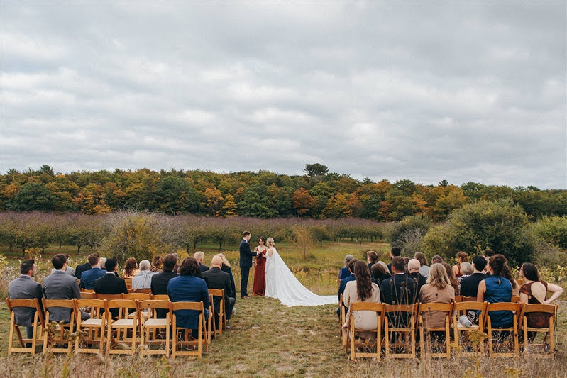 Wide view of October wedding outdoor ceremony with guests sitting in natural wood folding chairs at Cherry Basket Farm. 